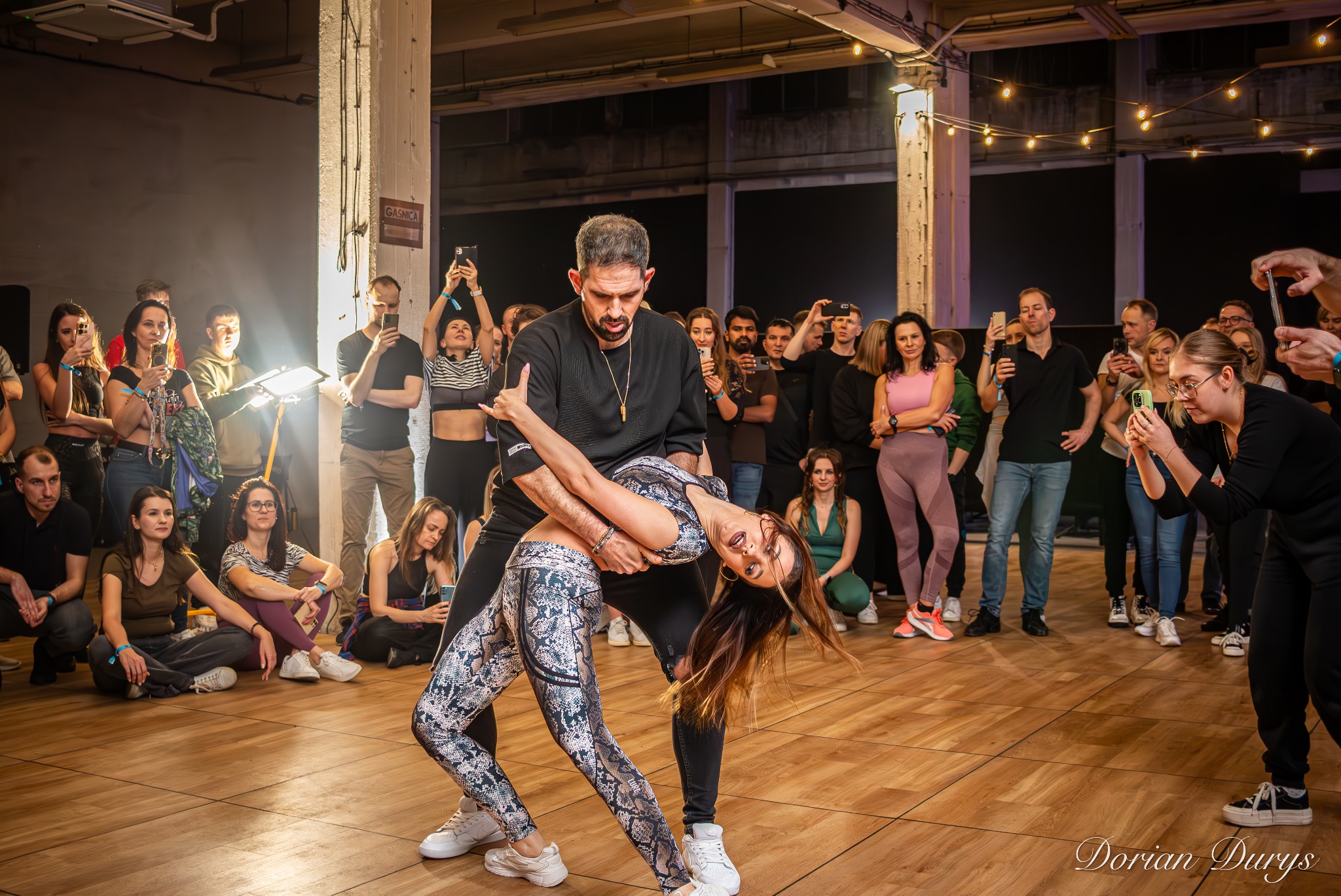 Para tańczy taniec towarzyski, kobieta w odchyleniu do tyłu, otoczeni przez widzów filmujących występ telefonami, w tle industrialna przestrzeń.