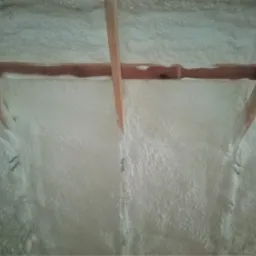 Poddasze ocieplone białą pianką natryskową, widoczne drewniane elementy konstrukcji dachu, gdzieniegdzie wystające metalowe elementy mocujące.