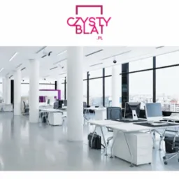 Jasne, przestronne biuro z białymi biurkami, krzesłami i kolumnami, dużą ilością naturalnego światła wpadającego przez okna, z widokiem na miasto w tle oraz logo firmy w górnej części kadru.