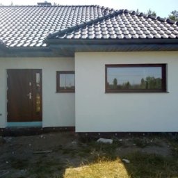Dam bud - Nowo otynkowany dom jednorodzinny z ciemnobrązową stolarką okienną i drzwiową, czarną dachówką i widocznym ociepleniem fundamentów, teren wokół domu jeszcze niezagospodarowany.