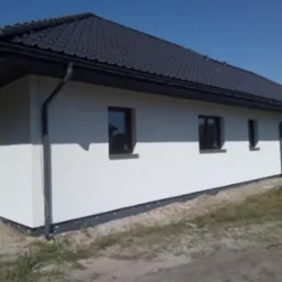 Wykończona elewacja domu jednorodzinnego z czarną dachówką i orynnowaniem, widok na narożnik budynku i fragment ściany z oknami, teren wokół budynku częściowo zagospodarowany.