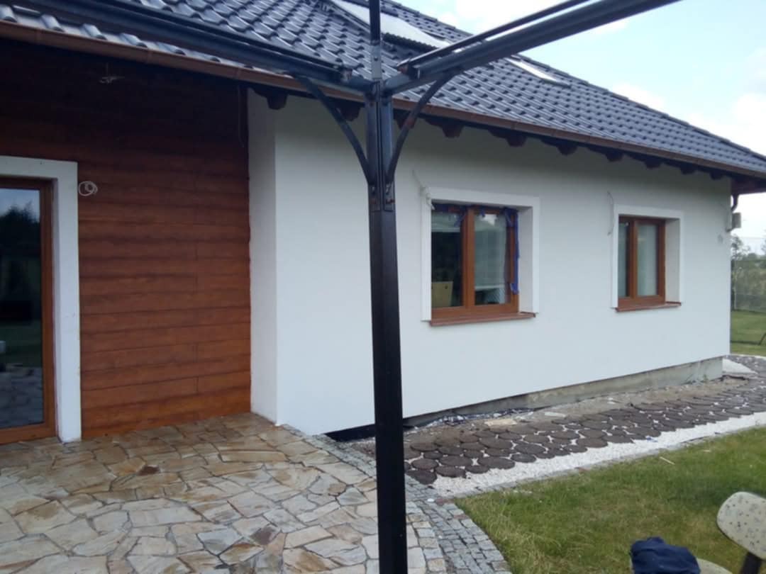 Wykończona elewacja domu jednorodzinnego z białym tynkiem i drewnopodobną okładziną, widoczne okna z brązowymi ramami, kamienny chodnik i czarna pergola.