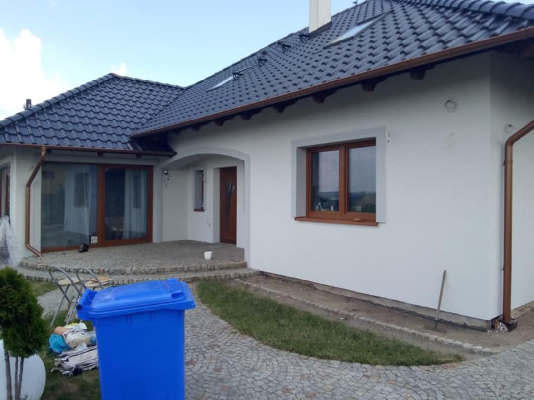 Biała elewacja domu z brązowymi oknami i ciemną dachówką, widoczny niebieski kosz na śmieci i drobne prace wykończeniowe wokół budynku.