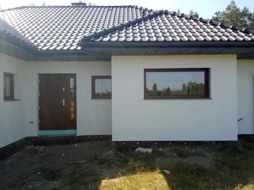 Nowo otynkowany dom jednorodzinny z ciemnobrązową stolarką okienną i drzwiową, czarną dachówką i widocznym ociepleniem fundamentów, teren wokół domu jeszcze niezagospodarowany.