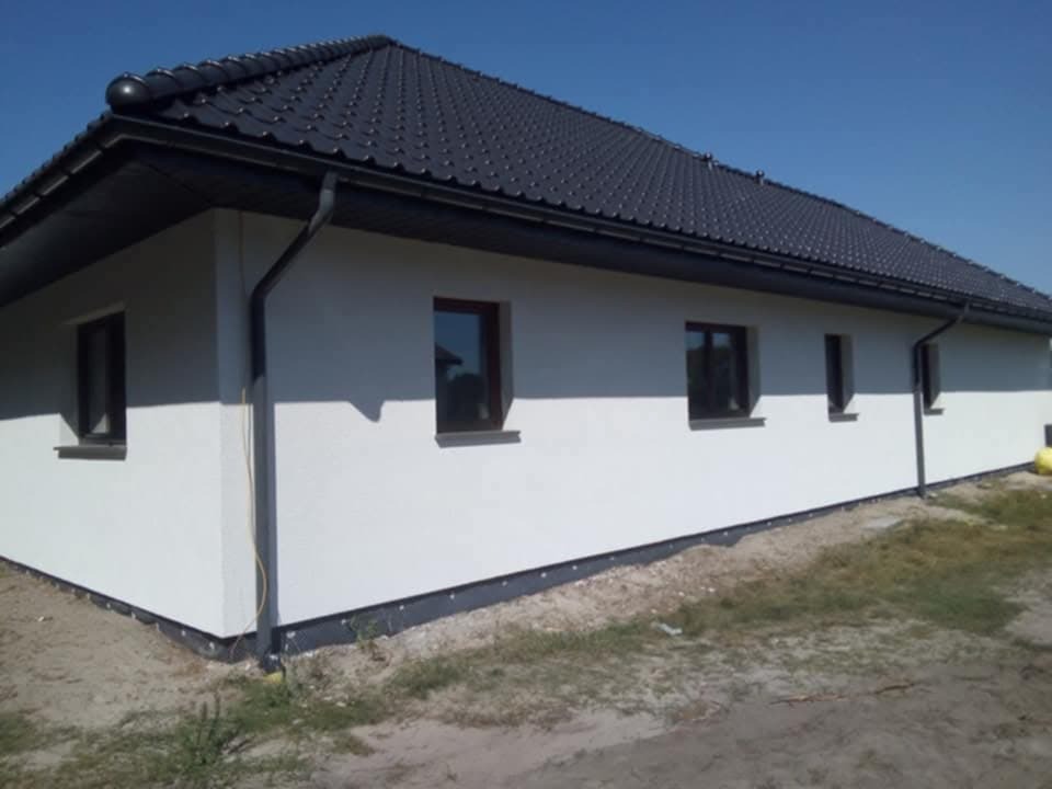 Wykończona elewacja domu jednorodzinnego z czarną dachówką i orynnowaniem, widok na narożnik budynku i fragment ściany z oknami, teren wokół budynku częściowo zagospodarowany.