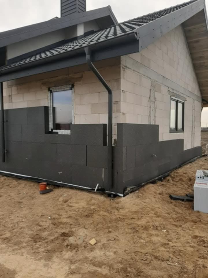 Ocieplanie elewacji domu jednorodzinnego z bloczków betonowych grafitowym styropianem, widoczne fragmenty niezabezpieczonej ściany, rynna i okno.