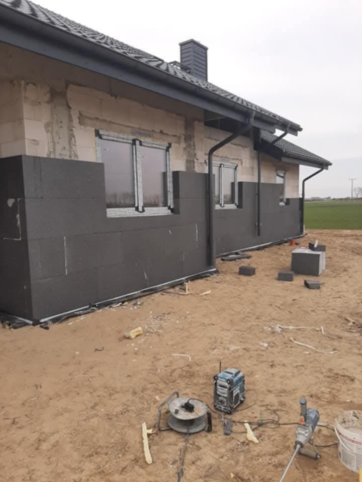 Ocieplanie elewacji domu jednorodzinnego: fragment ściany z zamontowaną izolacją styropianową, widoczne narzędzia budowlane na piaszczystym podłożu.