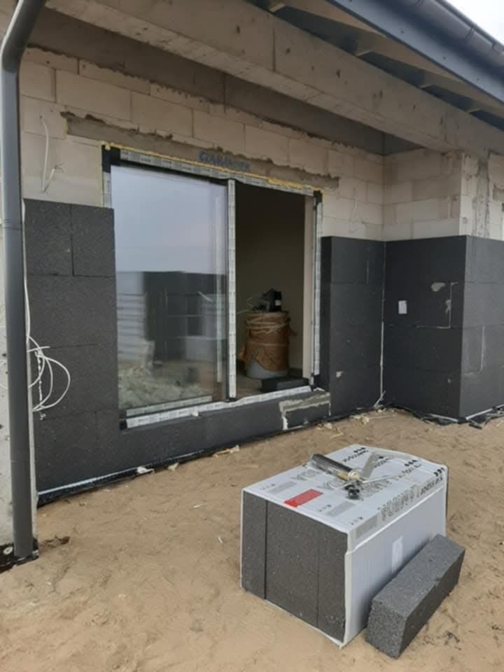 Montaż czarnych płyt izolacyjnych wokół okna przesuwnego w surowym stanie, widoczny paczka materiałów i narzędzia na piasku.