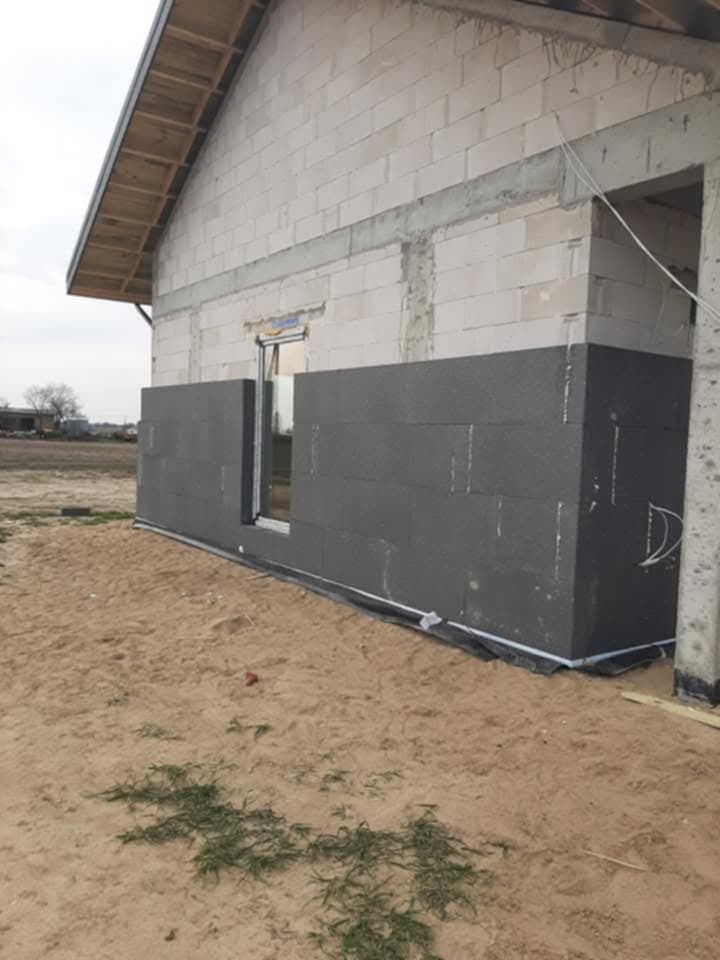 Ocieplanie ściany budynku szarym styropianem, widoczne bloczki betonowe i okno w stanie surowym.