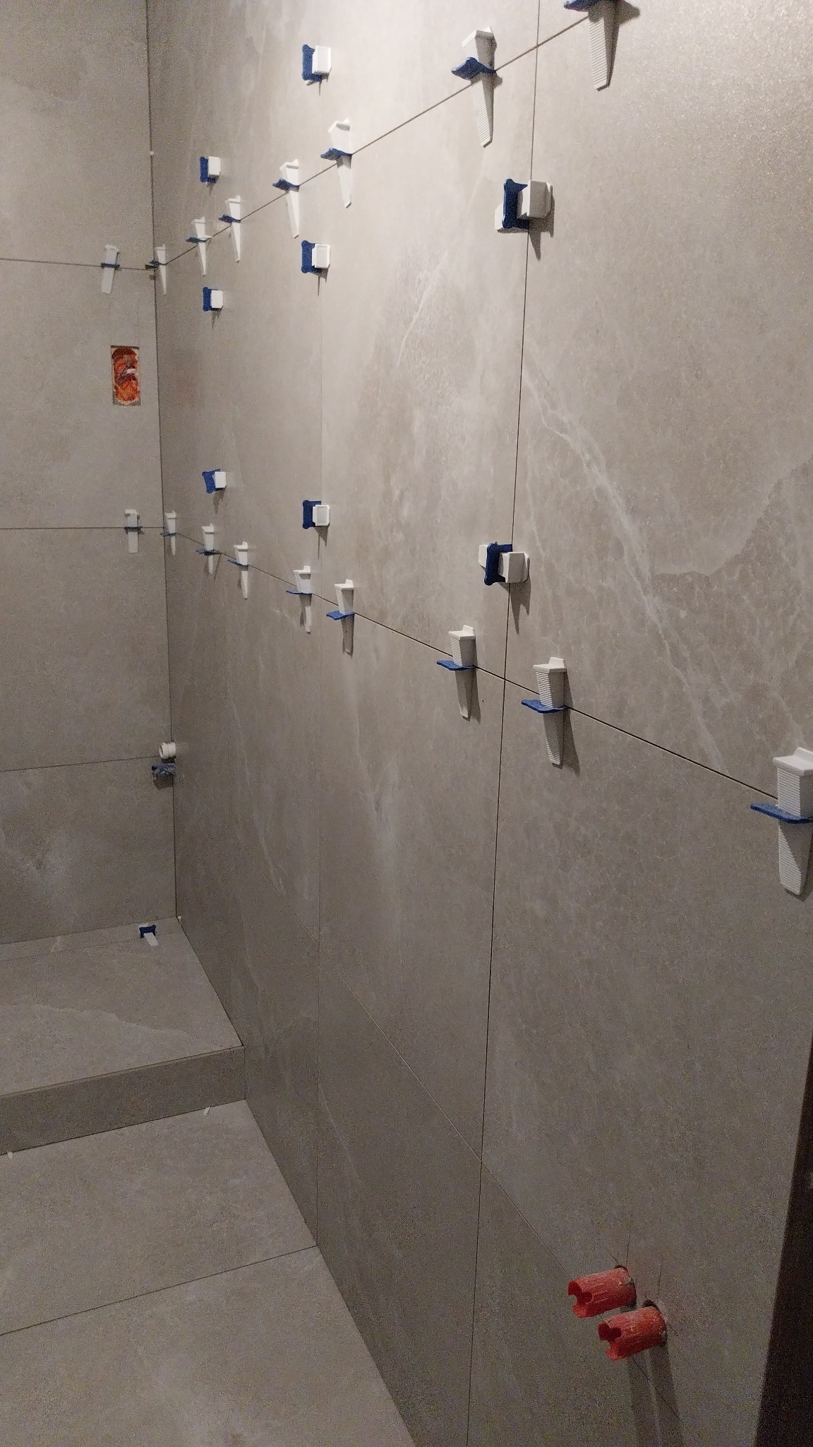 Proces układania płytek ceramicznych na ścianie łazienki z użyciem systemu poziomowania płytek, widoczne sznurki i klipsy dystansowe, z czerwonymi zaślepkami na rurach.