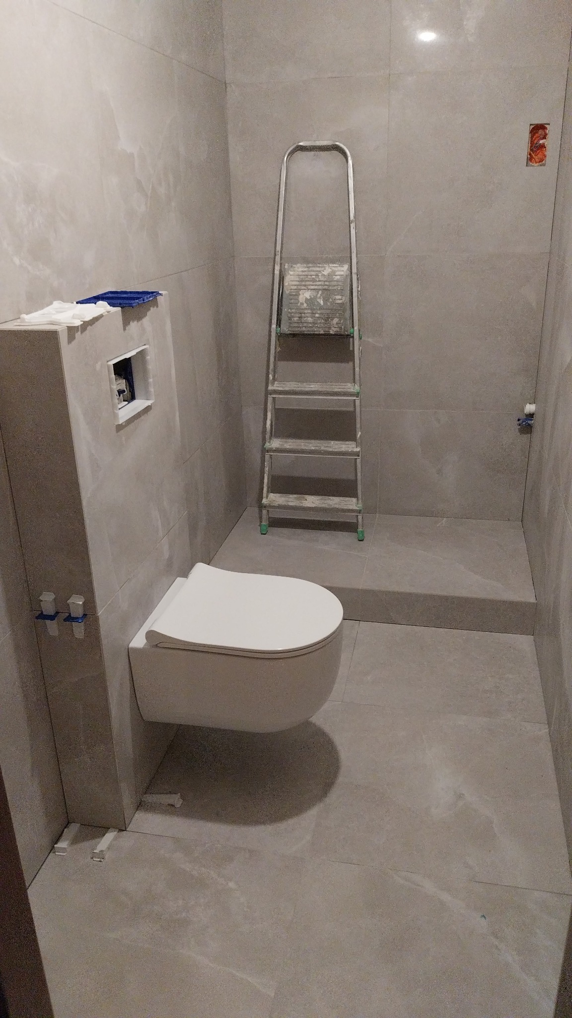 Podwieszana miska WC zamontowana w nowoczesnej łazience wykończonej płytkami imitującymi marmur; widoczna drabina malarska i elementy instalacji.