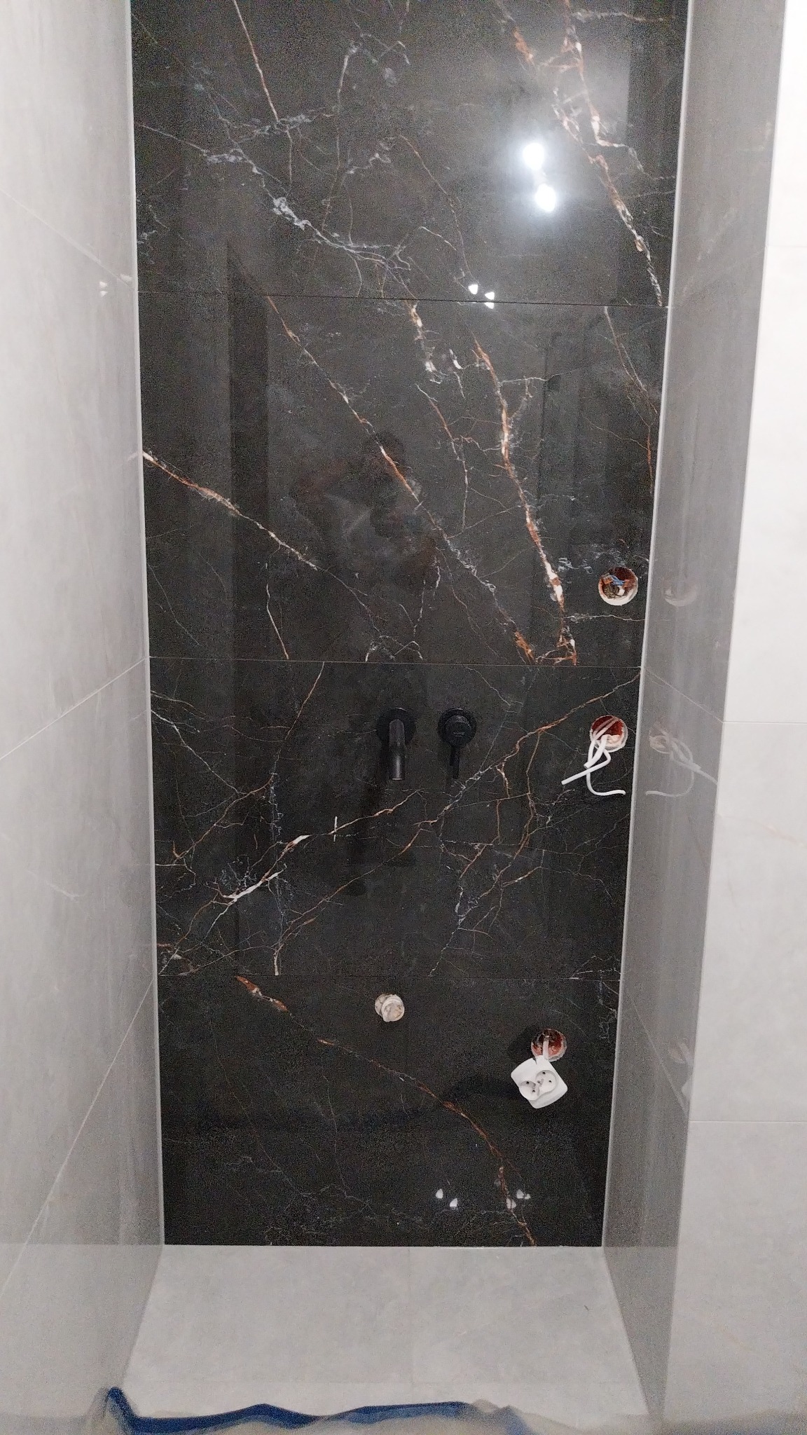 Częściowo wykończona ściana w łazience z czarnych płytek imitujących marmur ze złotymi żyłami, widoczne czarne baterie i wystające kable elektryczne.