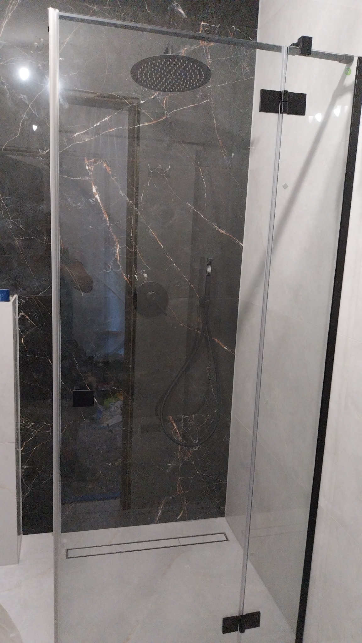 Kabina prysznicowa typu walk-in z czarnymi elementami i deszczownicą na tle ściany wyłożonej płytkami imitującymi czarny marmur z jasnymi żyłami.