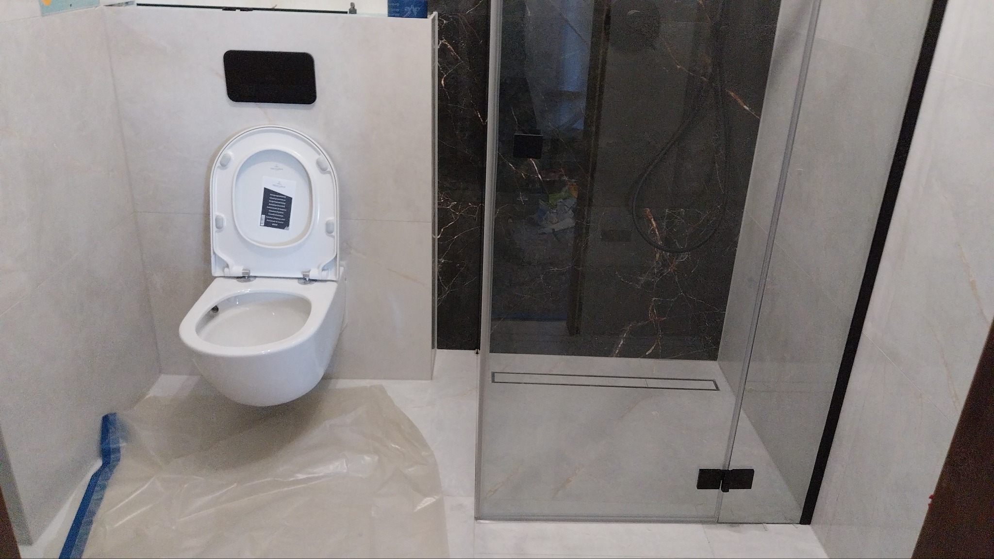 Nowoczesna łazienka z podwieszaną toaletą, kabiną prysznicową typu walk-in z czarnym wykończeniem i marmurowymi płytkami na ścianach, zabezpieczona folią malarską.