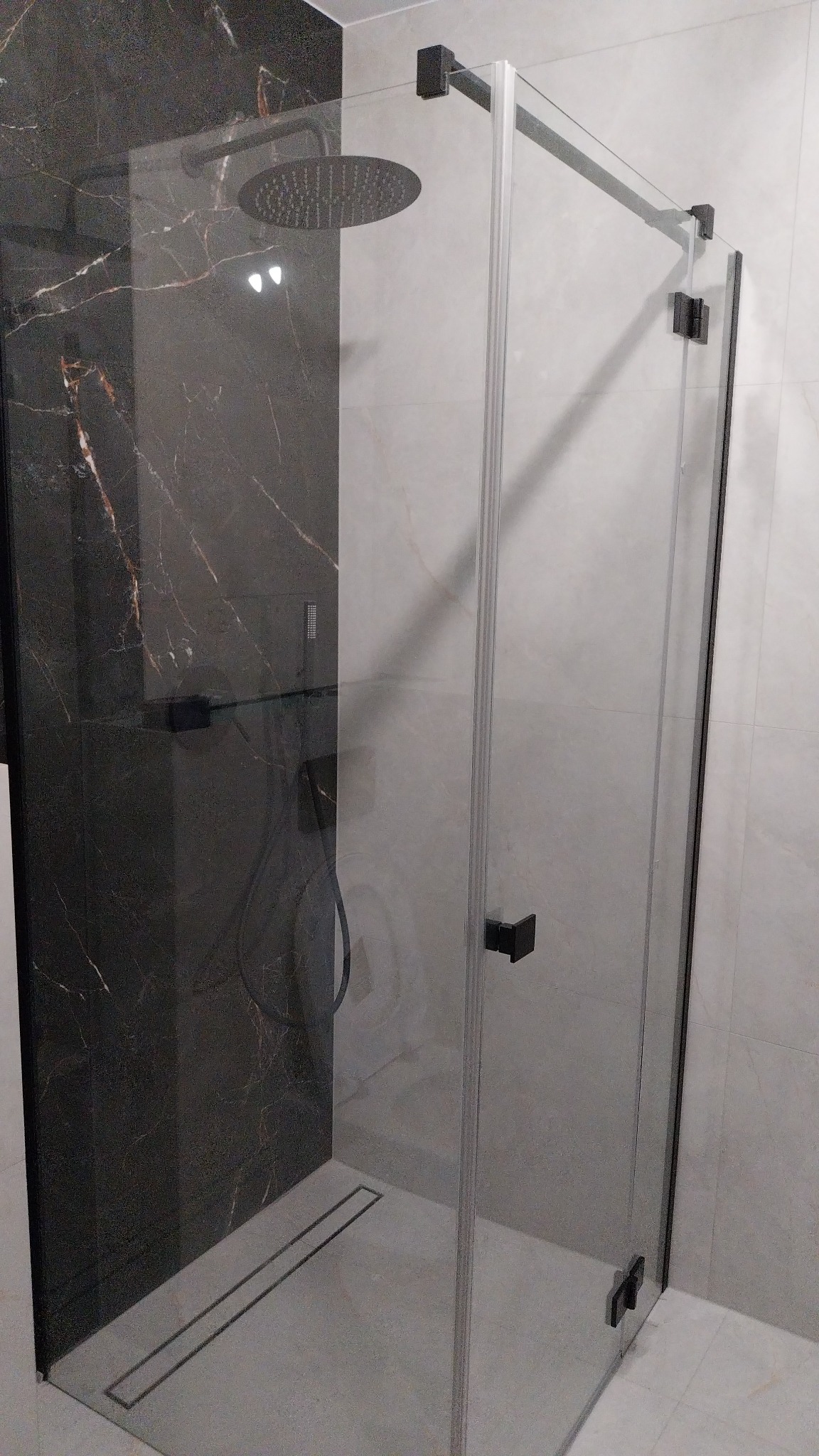 Nowoczesna kabina prysznicowa z czarnymi elementami, deszczownicą i odpływem liniowym w posadzce, wykończona płytkami imitującymi marmur.