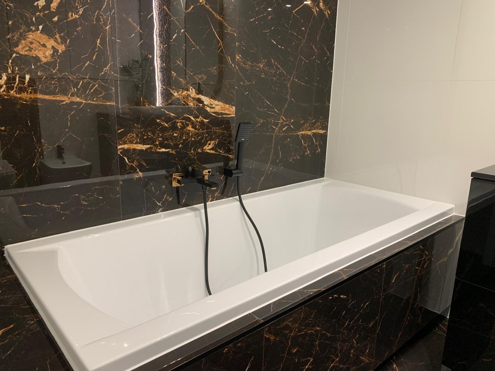 Biała wanna obudowana czarnym marmurem z złotymi żyłami, czarna bateria wannowa z elementami w kolorze złota, ściana wyłożona płytkami imitującymi czarny marmur, oświetlenie LED odbijające się...