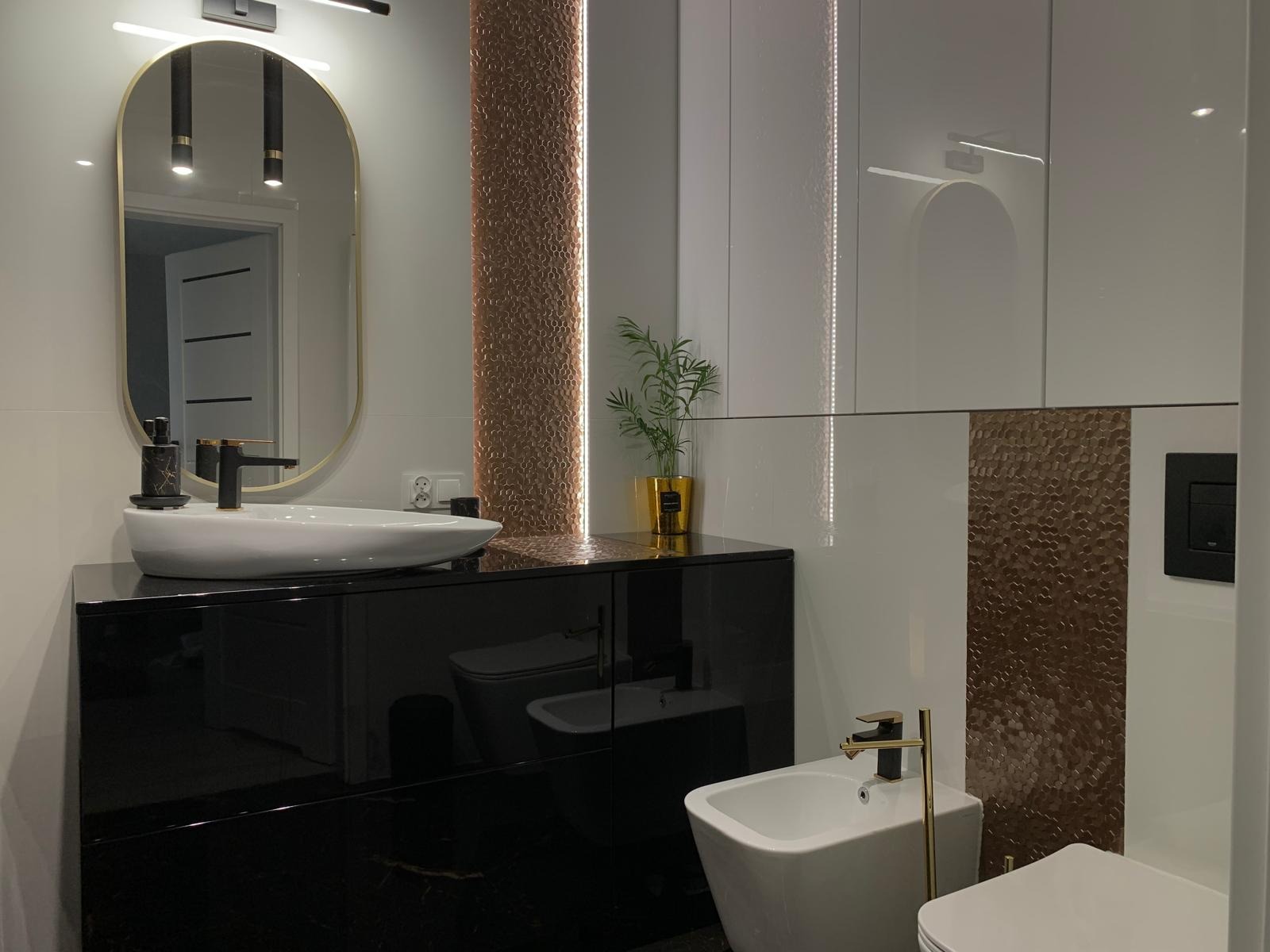 Nowoczesna łazienka z czarną szafką pod umywalkę, owalnym lustrem w złotej ramie, dekoracyjną mozaiką w odcieniach brązu i białą armaturą.