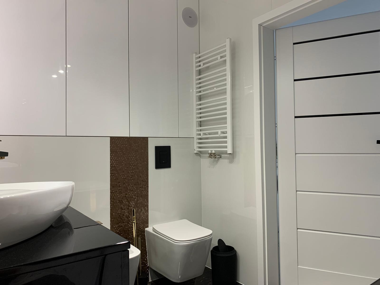 Nowoczesna łazienka z wiszącą miską WC, grzejnikiem drabinkowym, umywalką nablatową na czarnym blacie i szafkami w białym połysku.