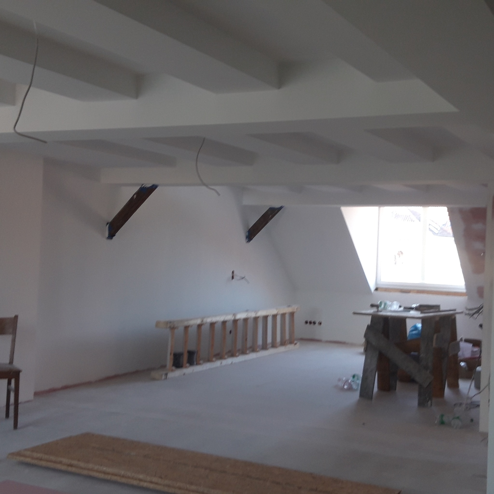 Poddasze w trakcie prac wykończeniowych: widoczne belki podtrzymujące dach, drewniana drabina oparta o ścianę, prowizoryczny stół roboczy i ślady po gładzi na ścianach.