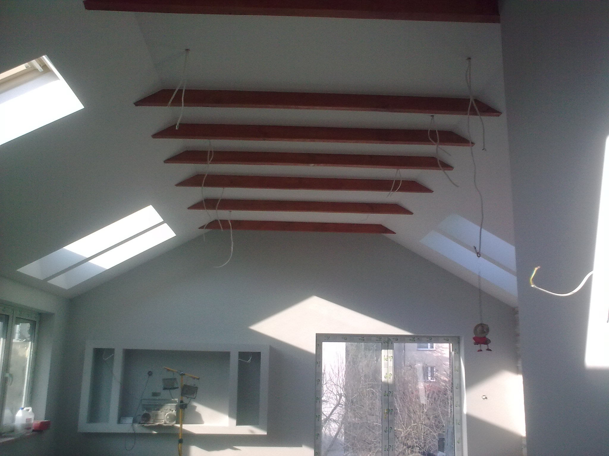 Poddasze w trakcie aranżacji z drewnianymi belkami pod sufitem, widoczne okna dachowe, lampa robocza i zabawka wisząca na kablu.