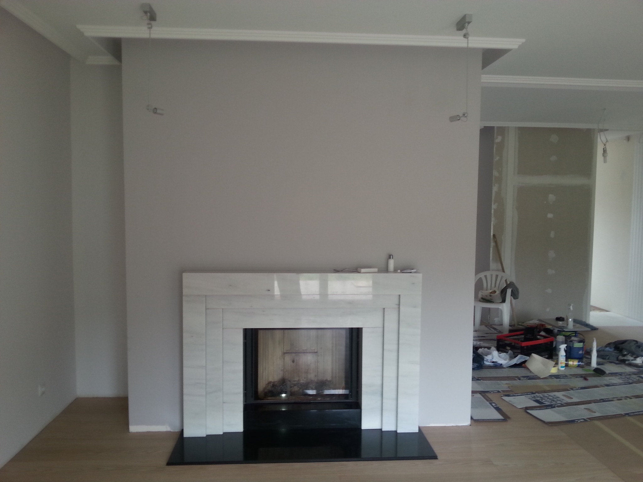Częściowo wykończony salon z marmurowym obramowaniem kominka i czarną podstawą, widoczne narzędzia i materiały budowlane w tle, ściany pomalowane na szaro, podwieszane oświetlenie sufitowe.