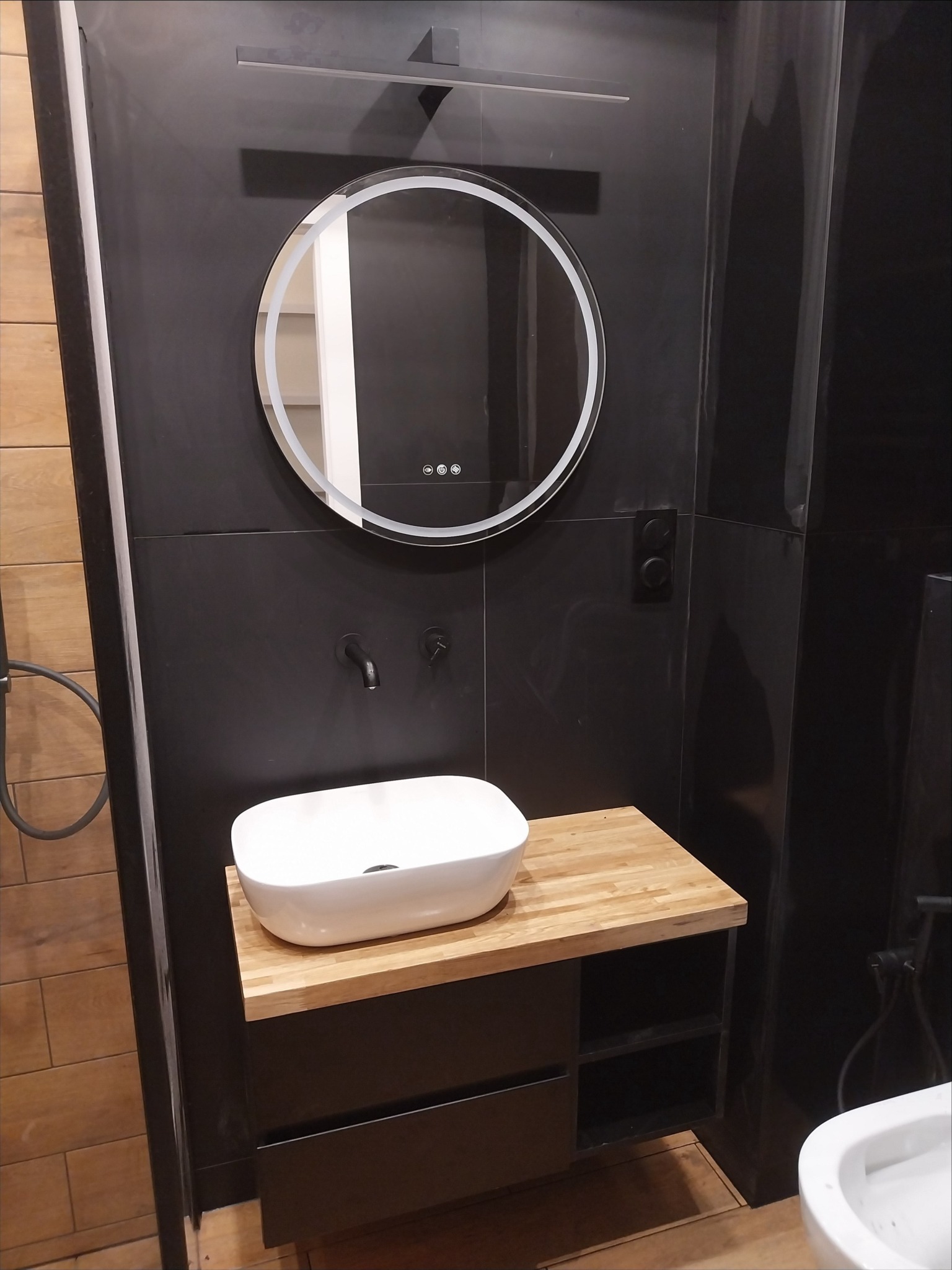 Nowoczesna łazienka z czarnymi płytkami, okrągłym lustrem z podświetleniem LED, białą umywalką nablatową na drewnianym blacie i czarną armaturą.