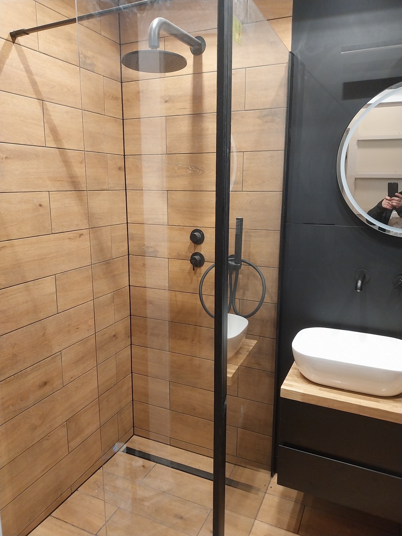 Nowoczesna kabina prysznicowa z drewnopodobnymi płytkami, czarną armaturą i okrągłym lustrem z podświetleniem LED. Umywalka nablatowa o owalnym kształcie na drewnianym blacie.
