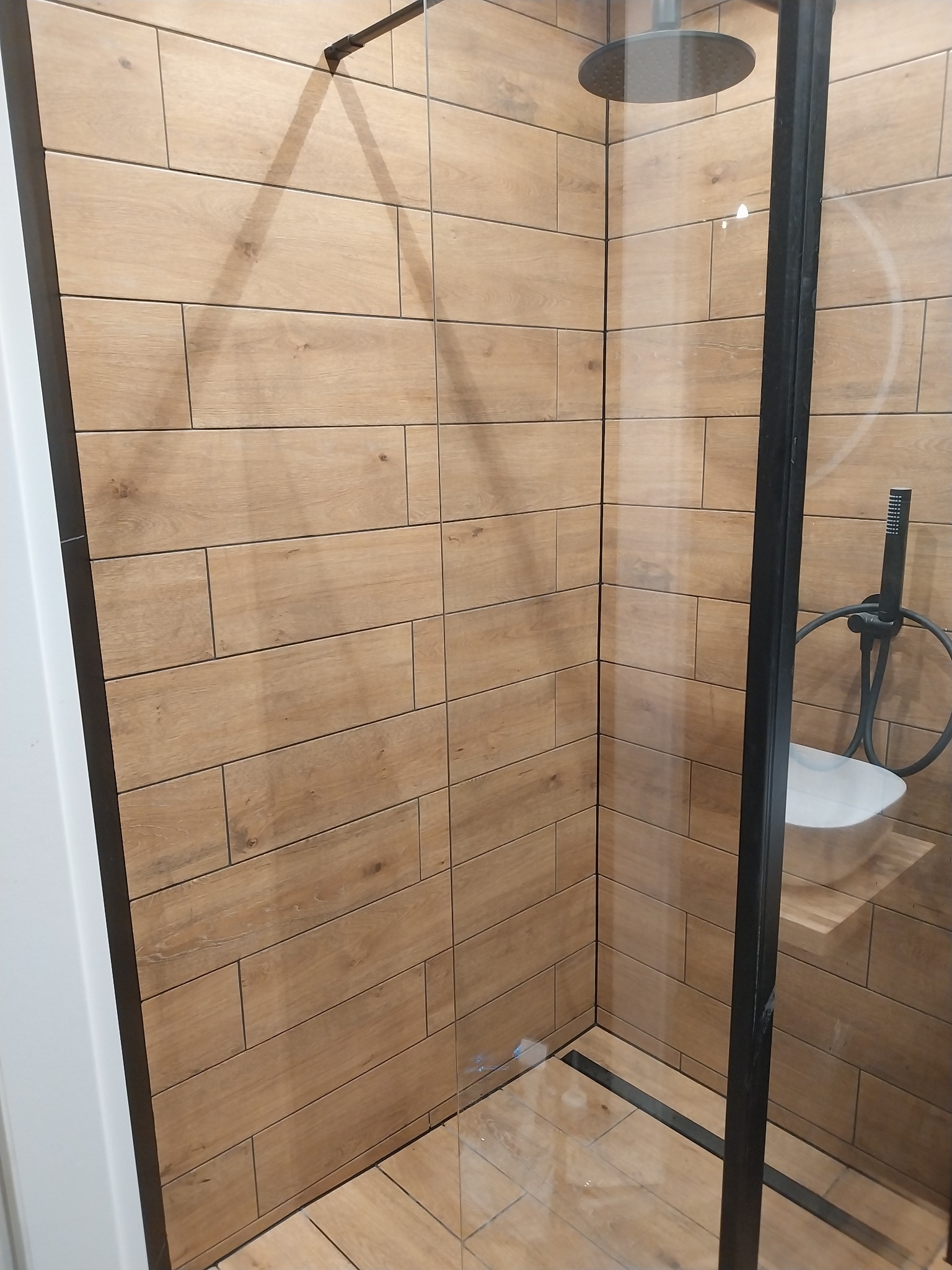 Kabina prysznicowa w Szczecinie z drewnopodobnymi płytkami na ścianach, deszczownicą i czarną armaturą, widok przez szklaną ściankę.