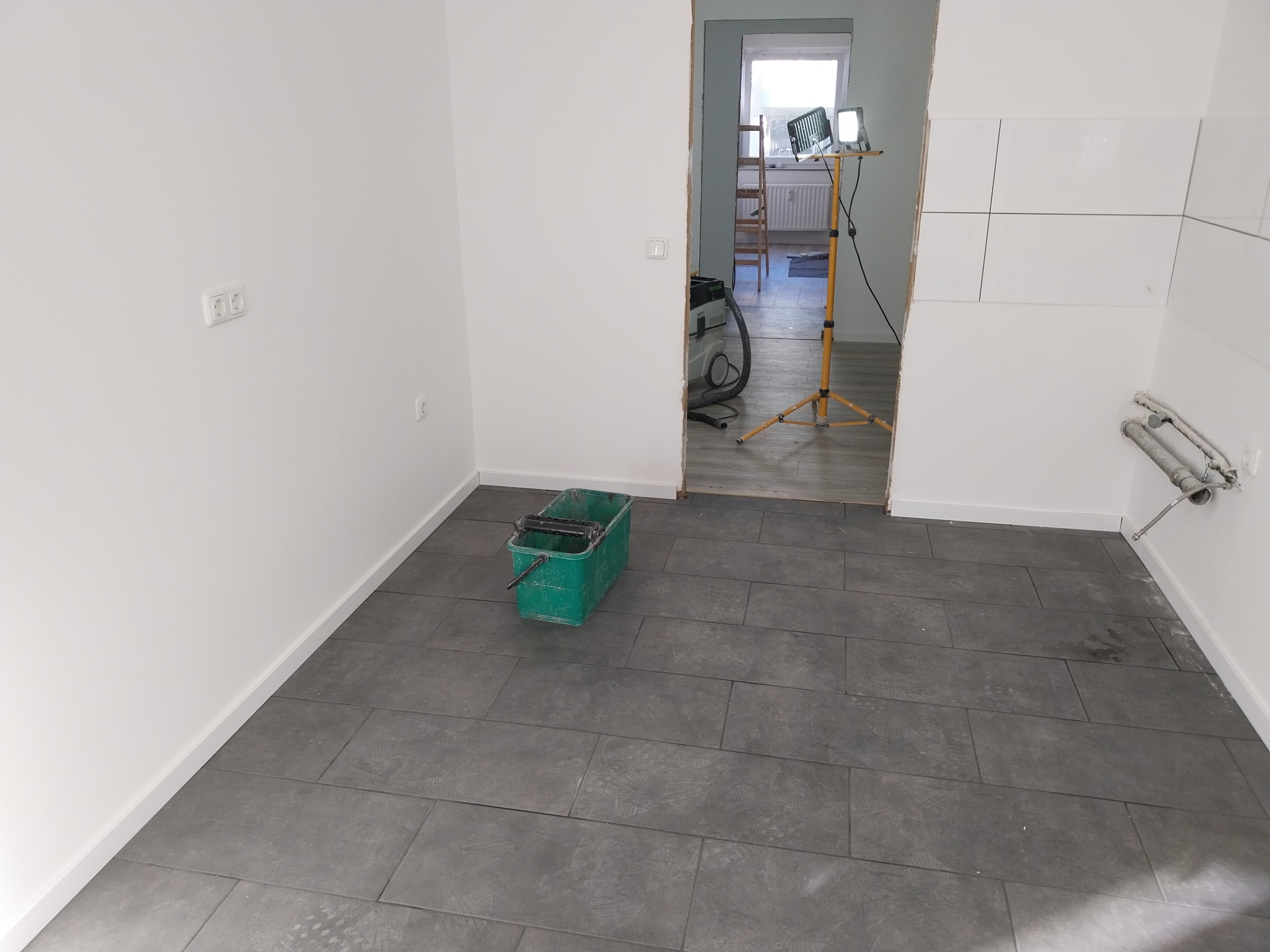 Wnętrze w trakcie remontu z szarymi płytkami na podłodze, białe ściany, wiadro z narzędziem malarskim, lampa budowlana i odkurzacz przemysłowy w przejściu do kolejnego pomieszczenia.