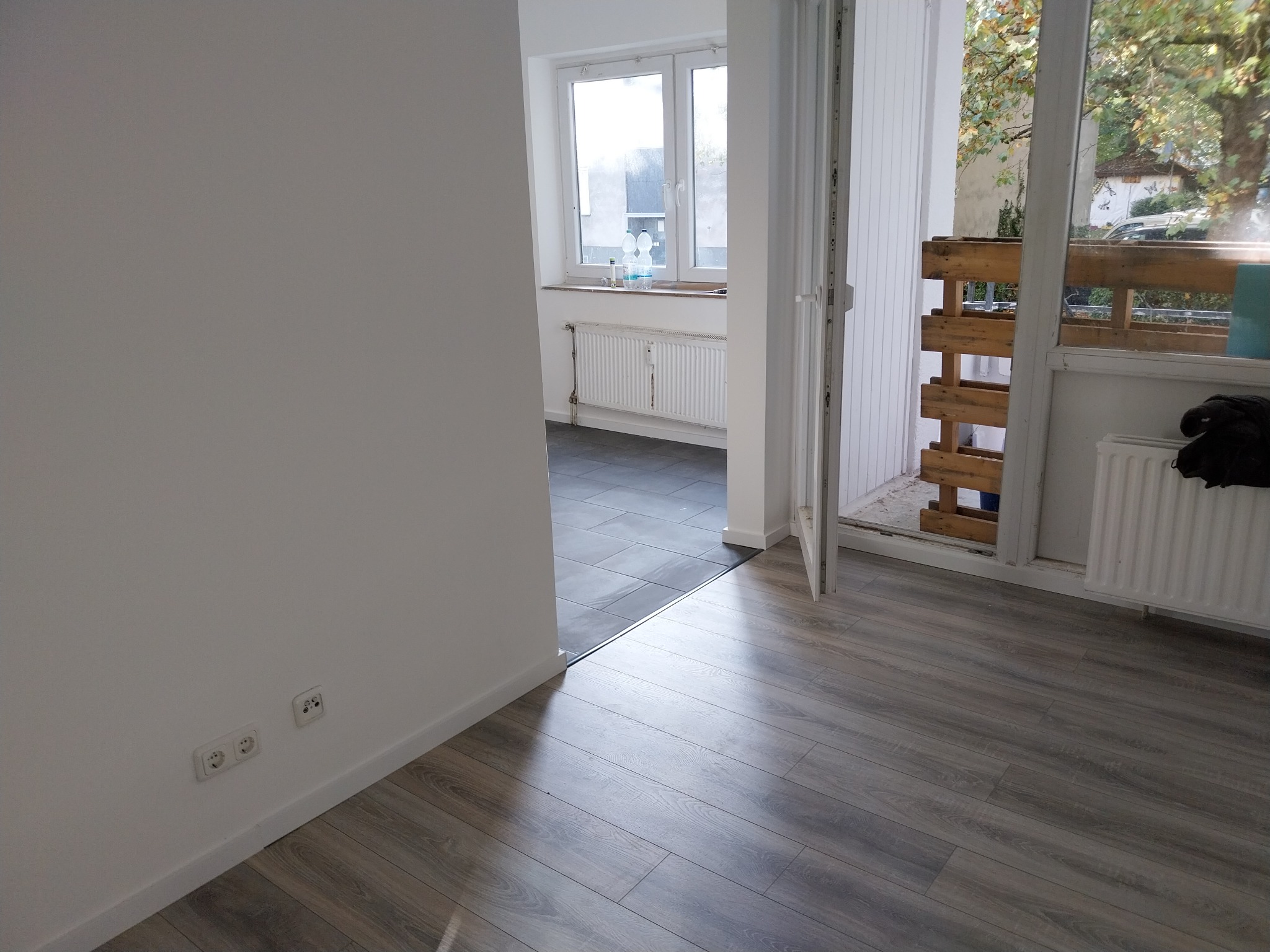 Pusta przestrzeń z szarą podłogą imitującą drewno, białe ściany z gniazdkami elektrycznymi, widok na pokój z grzejnikiem i oknem oraz balkon z drewnianą konstrukcją.