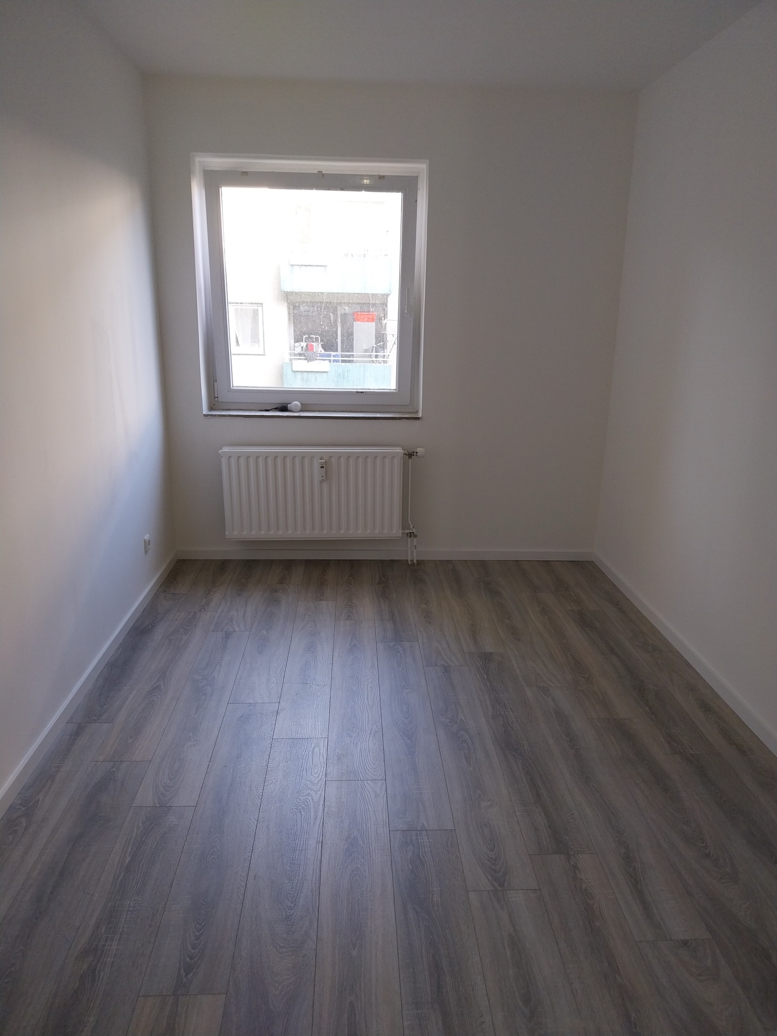Pusty pokój po remoncie z szarymi panelami podłogowymi, białymi ścianami i grzejnikiem pod oknem z widokiem na budynek.