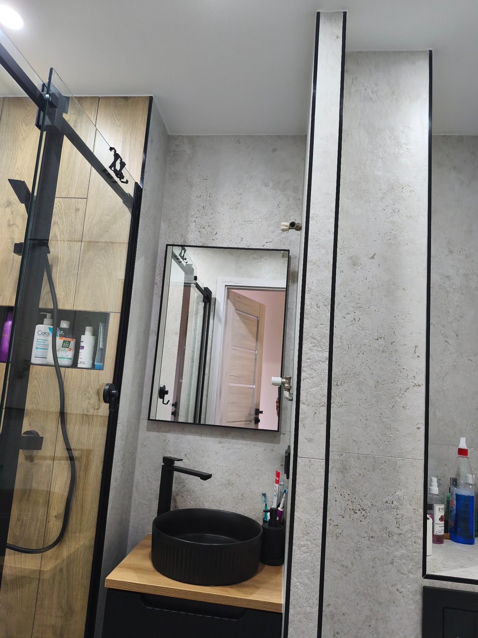 Nowoczesna łazienka z kabiną prysznicową typu walk-in, umywalką nablatową i lustrem w czarnej ramie. Ściany wykończone płytkami imitującymi beton.