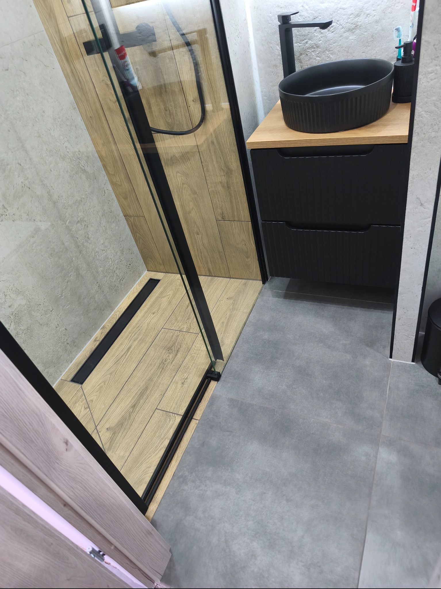 Nowoczesna łazienka z kabiną prysznicową typu walk-in z drewnopodobną podłogą, czarną armaturą i umywalką nablatową. Szare płytki na ścianach i podłodze.