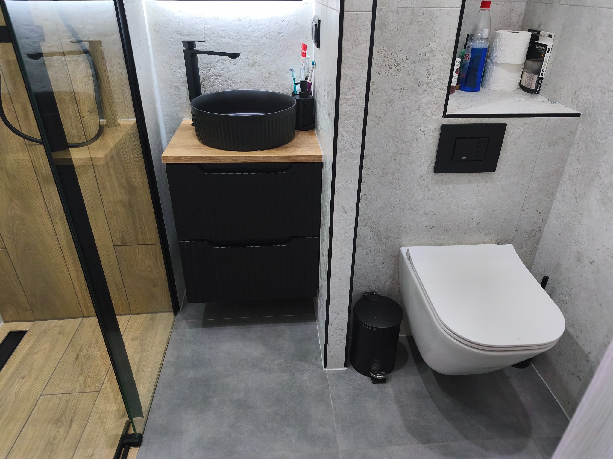 Nowoczesna łazienka z czarną umywalką nablatową i drewnianym blatem, szara płytka imitująca kamień, wisząca toaleta i kabina prysznicowa z drewnianym akcentem.