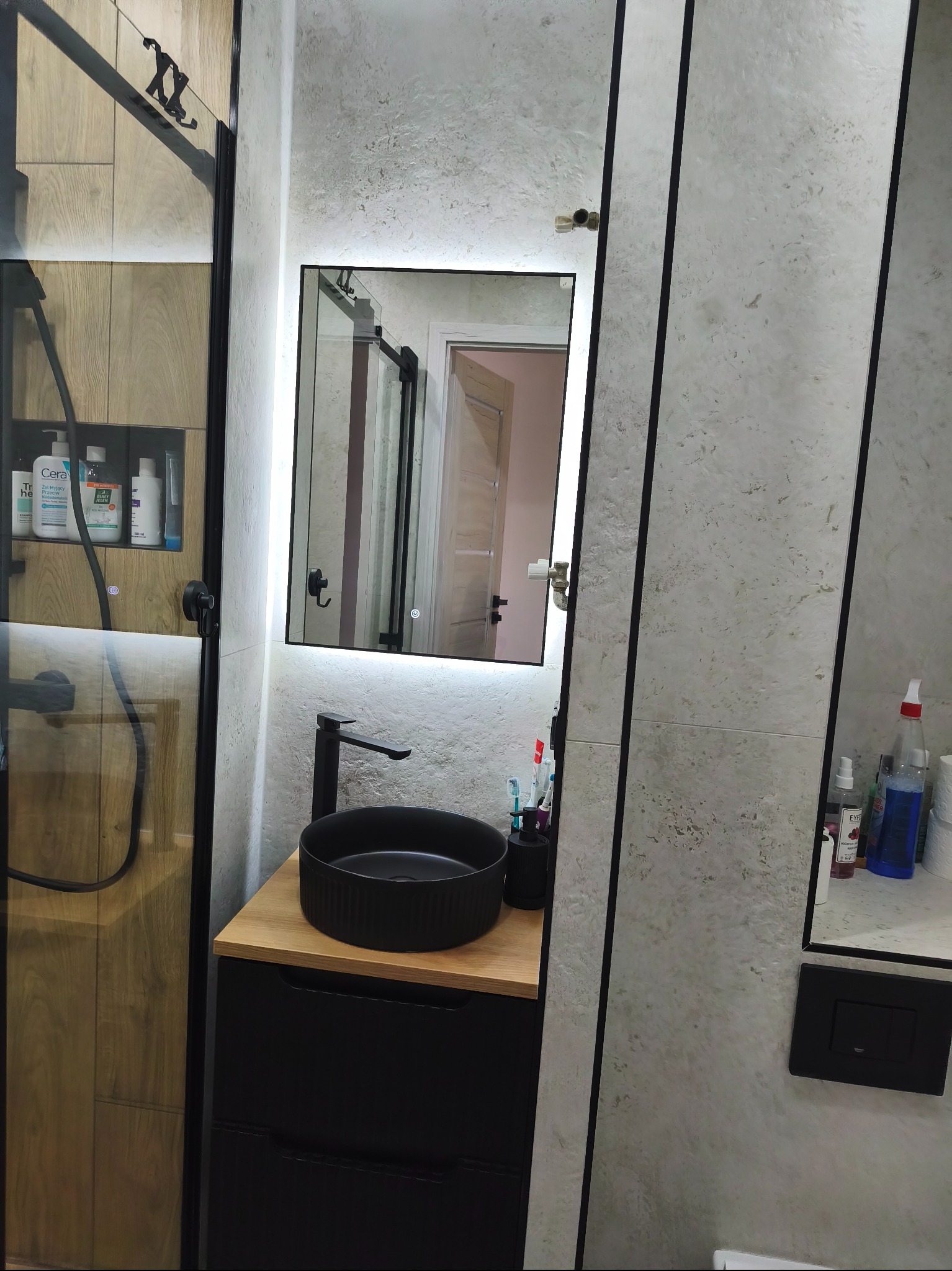Nowoczesna łazienka z czarną umywalką nablatową, podświetlanym lustrem i kabiną prysznicową z drewnianym akcentem. Minimalistyczny design, jasne płytki.
