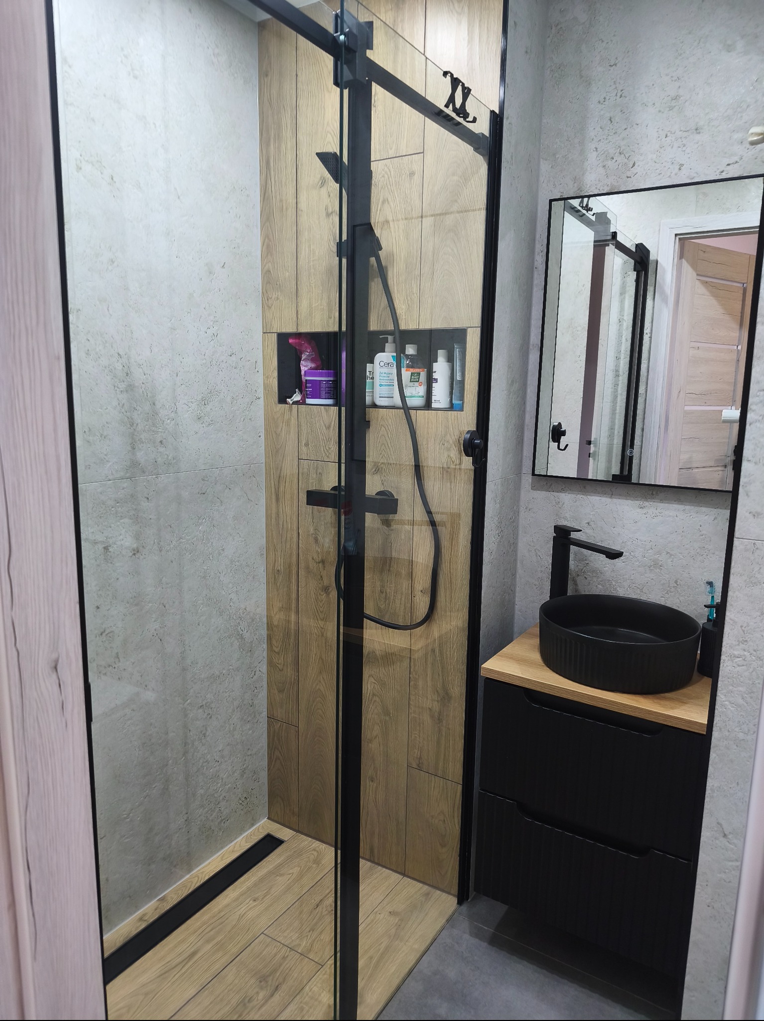 Nowoczesna łazienka z kabiną prysznicową typu walk-in, drewnopodobnymi płytkami i czarną armaturą. Umywalka nablatowa w kolorze czarnym. W tle lustro z podświetleniem.