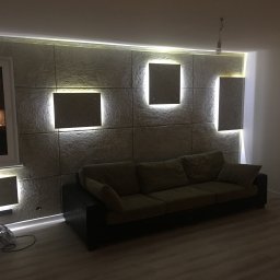 Naprawmy To - Szara sofa na tle dekoracyjnej ściany z paneli z podświetleniem LED, widoczny wentylator pod oknem.