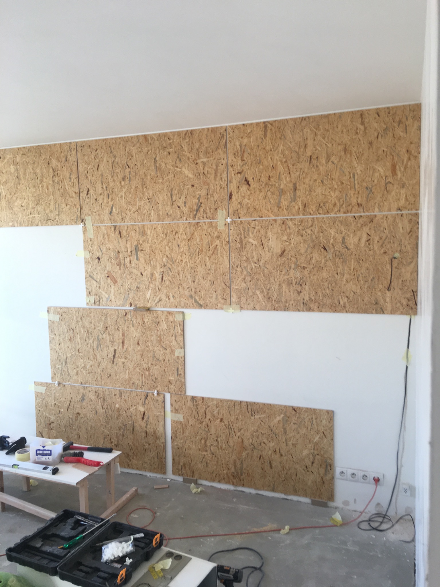 Płyty OSB mocowane do ściany taśmą malarską, widoczne narzędzia na stole i skrzynka narzędziowa, w tle gniazdka elektryczne.