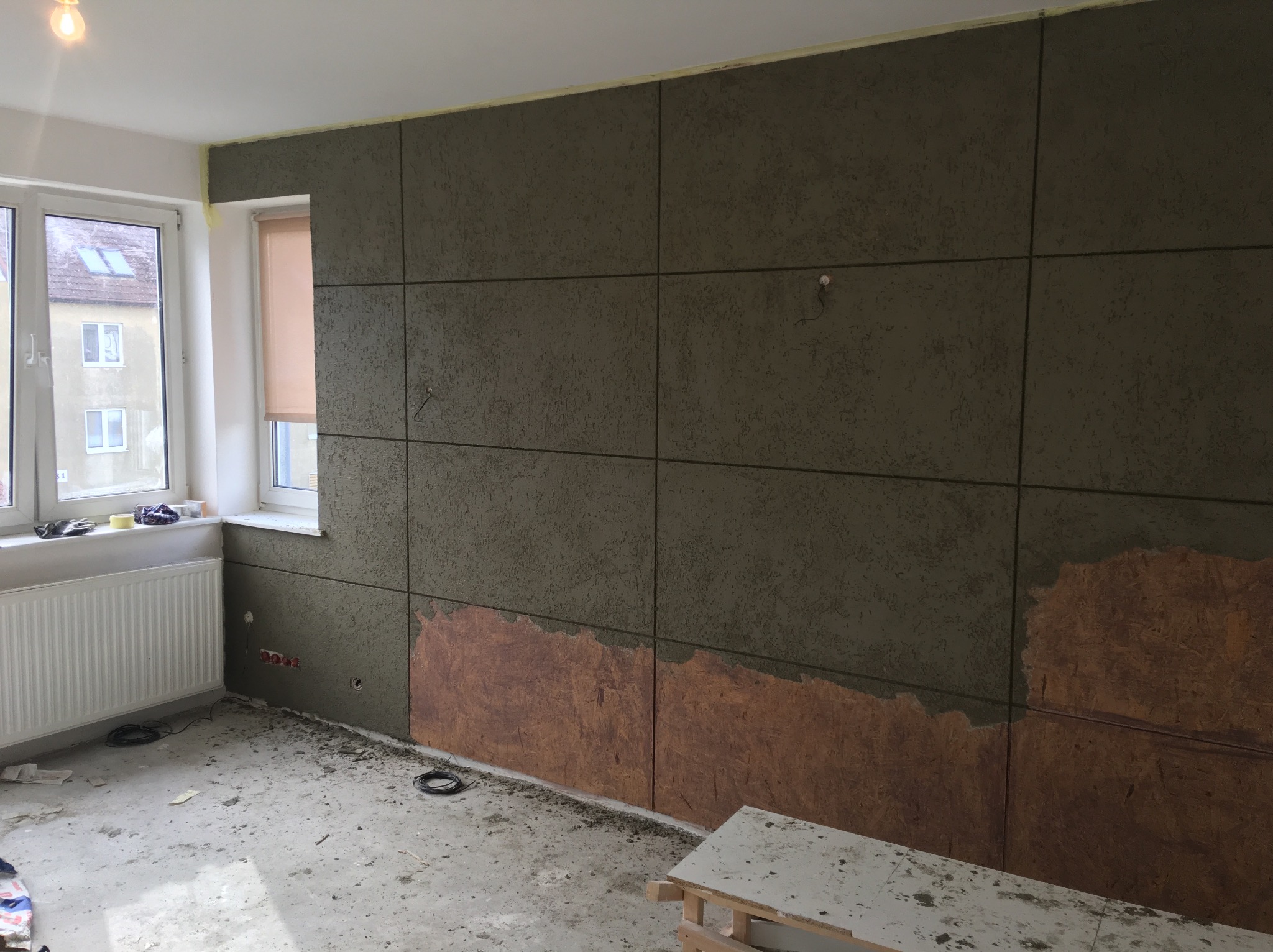 Montaż paneli ściennych w trakcie realizacji, widoczne fragmenty surowej ściany, okno z roletą, grzejnik i narzędzia na parapecie.