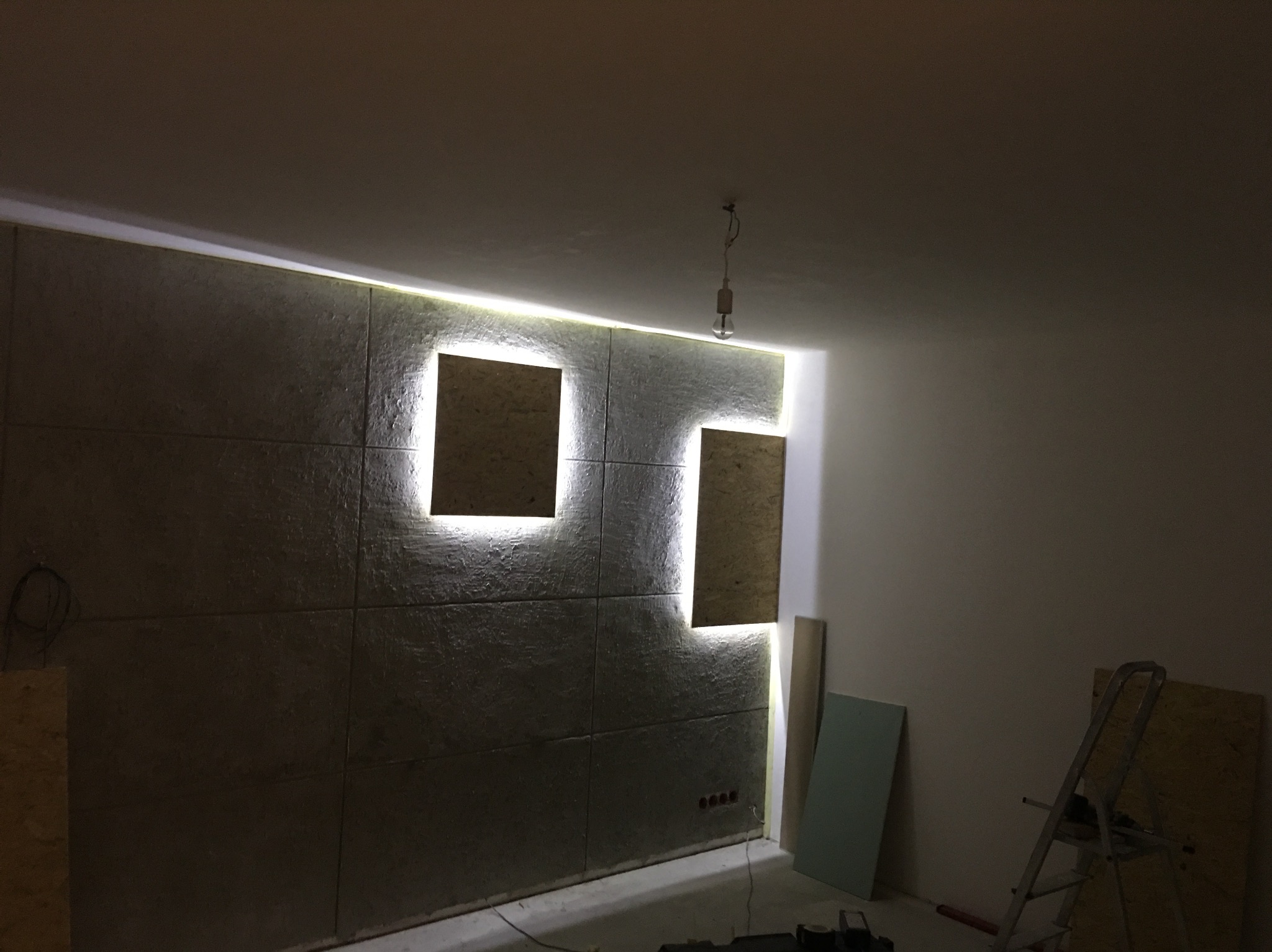 Ściana wyłożona panelami z oświetleniem LED w kształcie kwadratów, widoczne narzędzia i drabina, surowa żarówka zwisająca z sufitu, gniazdka elektryczne w ścianie