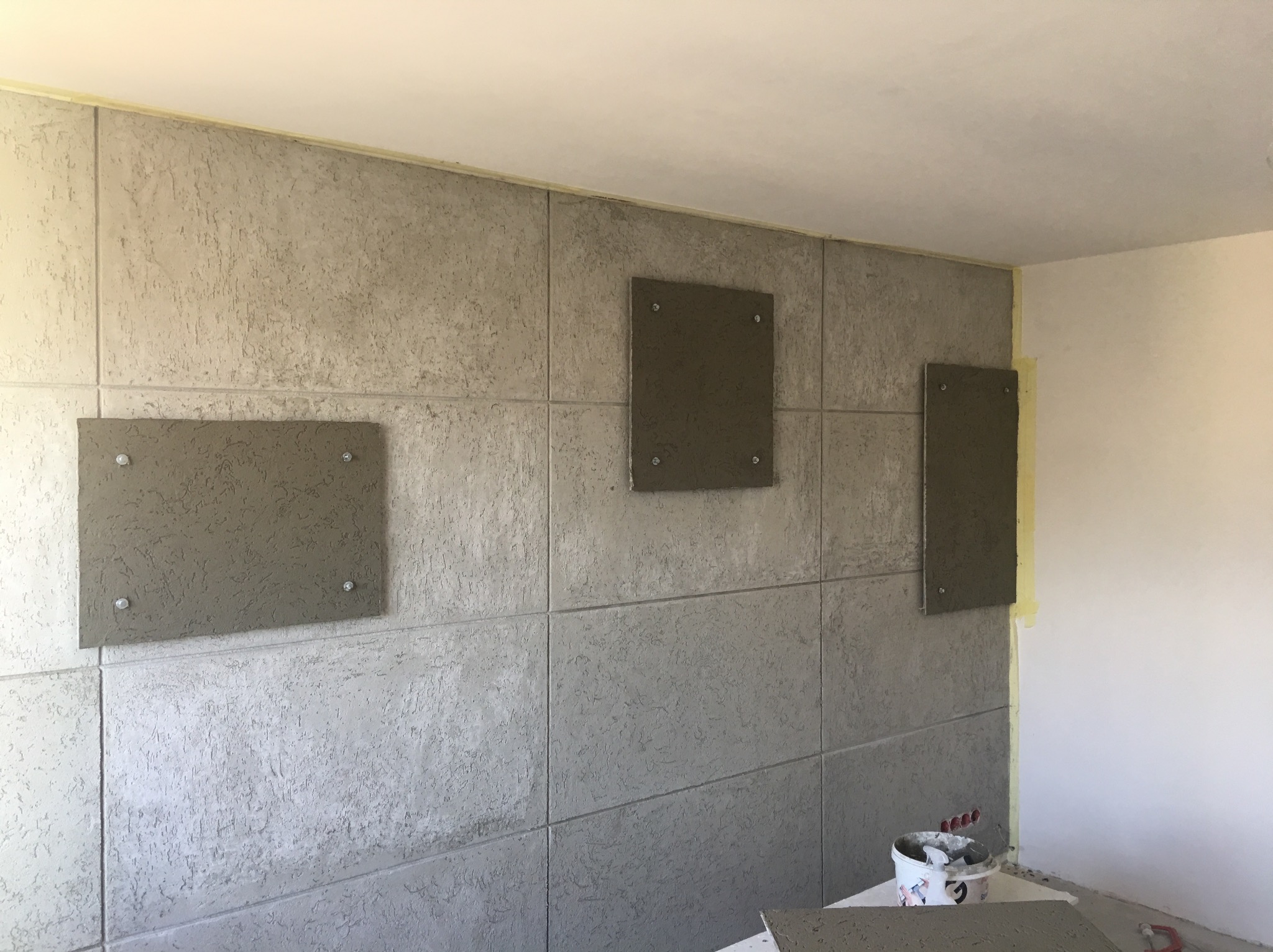 Montaż paneli ściennych o szorstkiej fakturze, trzy panele częściowo przymocowane do ściany za pomocą śrub, widoczna taśma malarska zabezpieczająca narożnik, wiadro z narzędziami w tle.