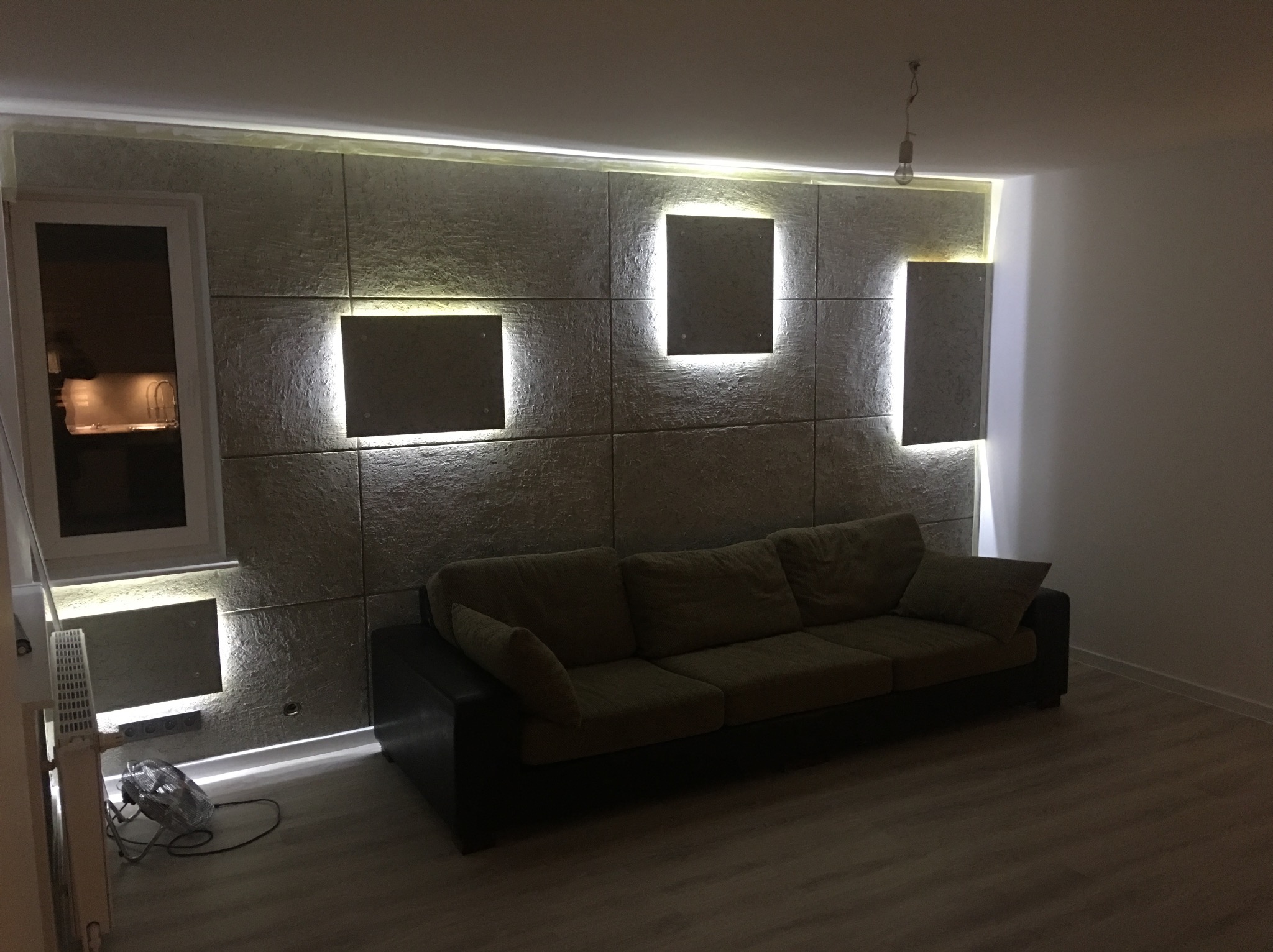 Szara sofa na tle dekoracyjnej ściany z paneli z podświetleniem LED, widoczny wentylator pod oknem.