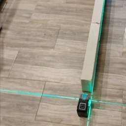 Damian Kielar - Poziomica laserowa wyznaczająca linię na podłodze z płytek ceramicznych, obok bloczek z betonu komórkowego.