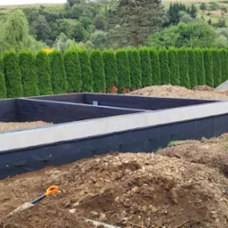 Wykop pod fundamenty budynku z izolacją przeciwwilgociową, widoczna łopata w ziemi i rząd tuj w tle.