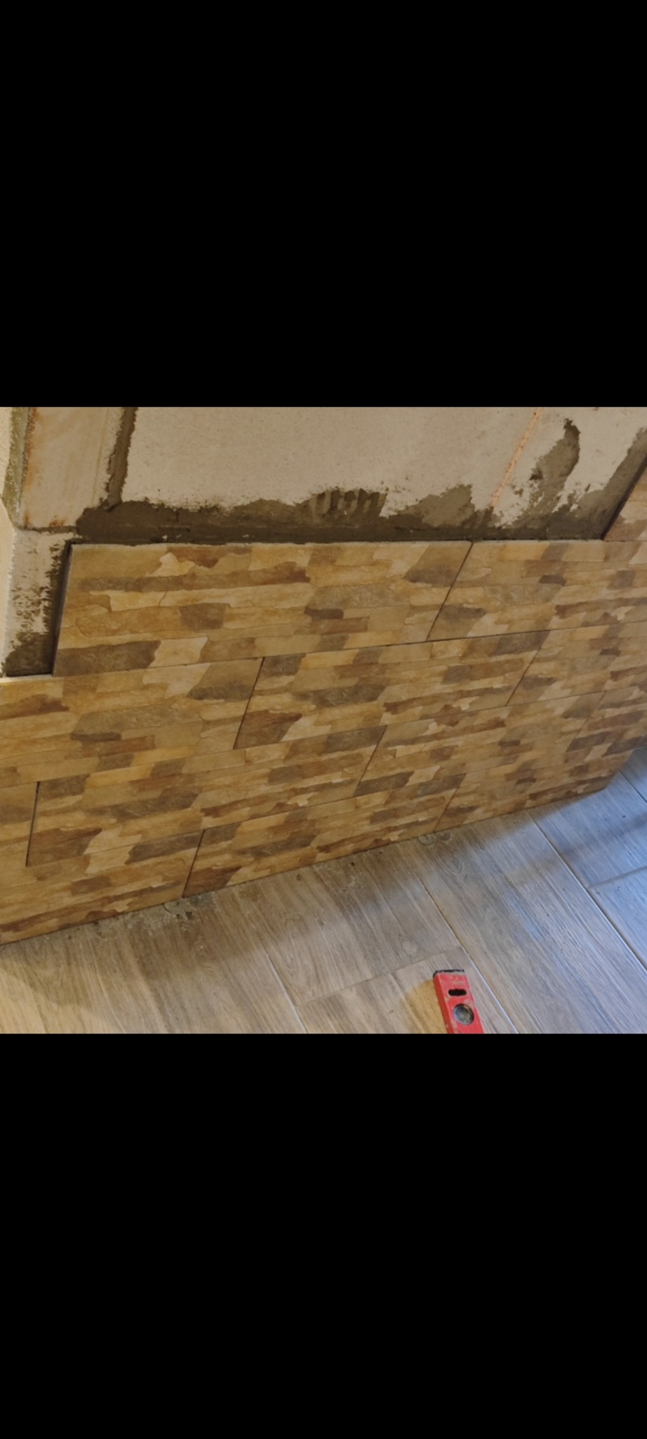 Fragment ściany z ułożoną dekoracyjną okładziną imitującą kamień, widoczne niedokończone prace oraz poziomica na podłodze.