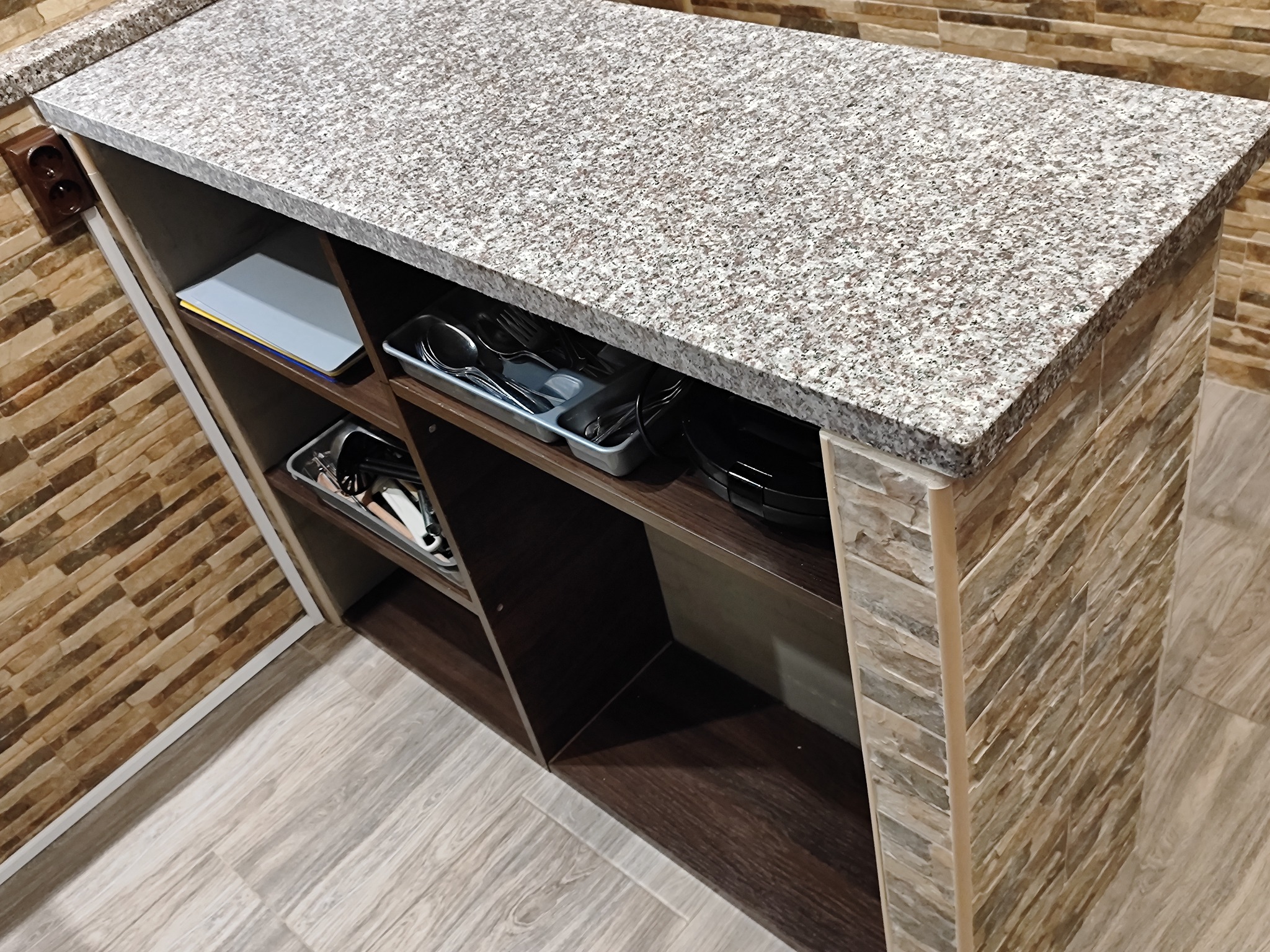 Wykończony fragment łazienki z blatem z jasnego granitu, półkami z ciemnego drewna i ścianą wyłożoną płytkami imitującymi kamień.