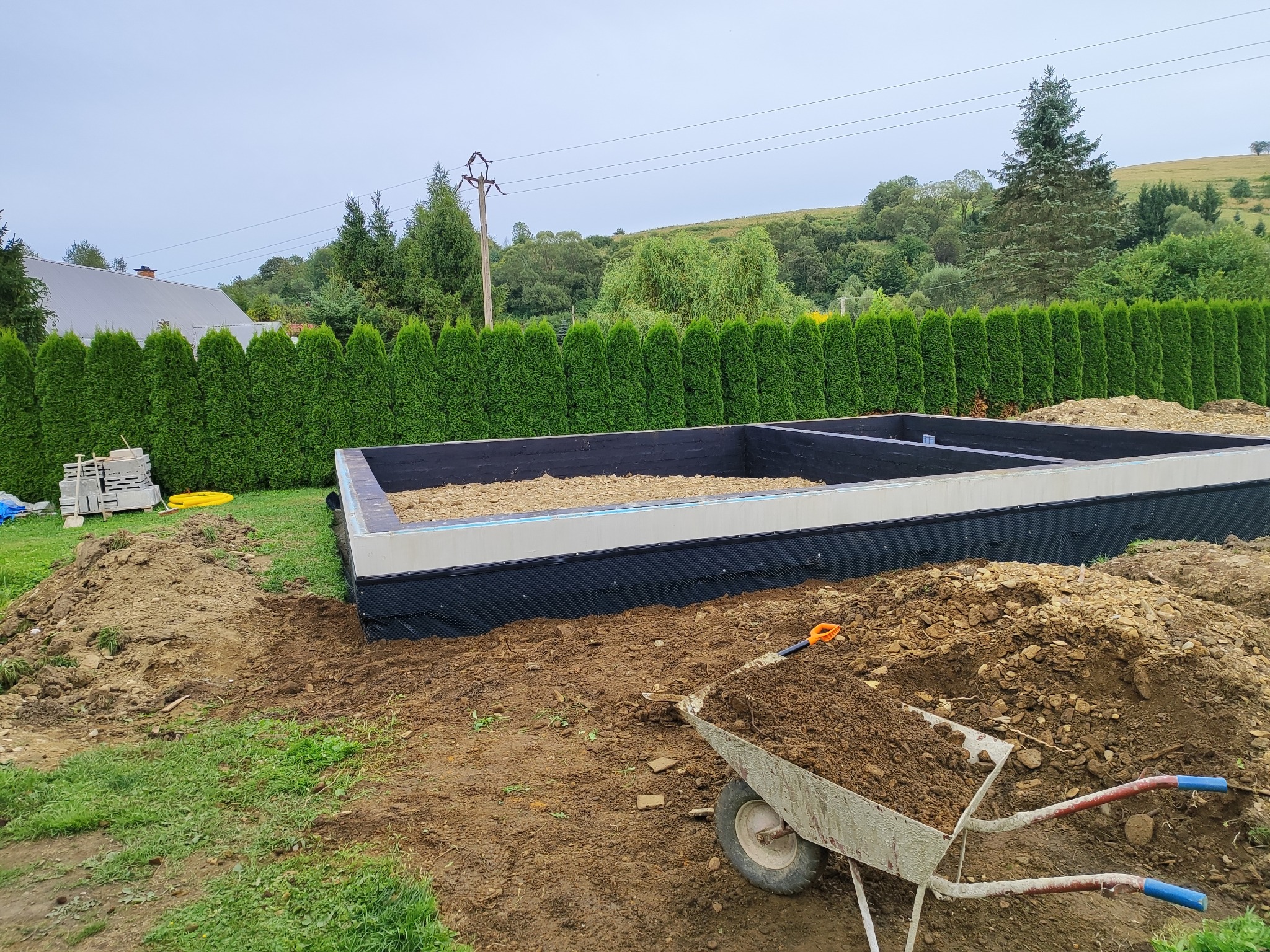 Fundament pod budynek w trakcie budowy, widoczna izolacja przeciwwilgociowa, taczka z ziemią na pierwszym planie, w tle zielony żywopłot i pagórkowaty krajobraz.