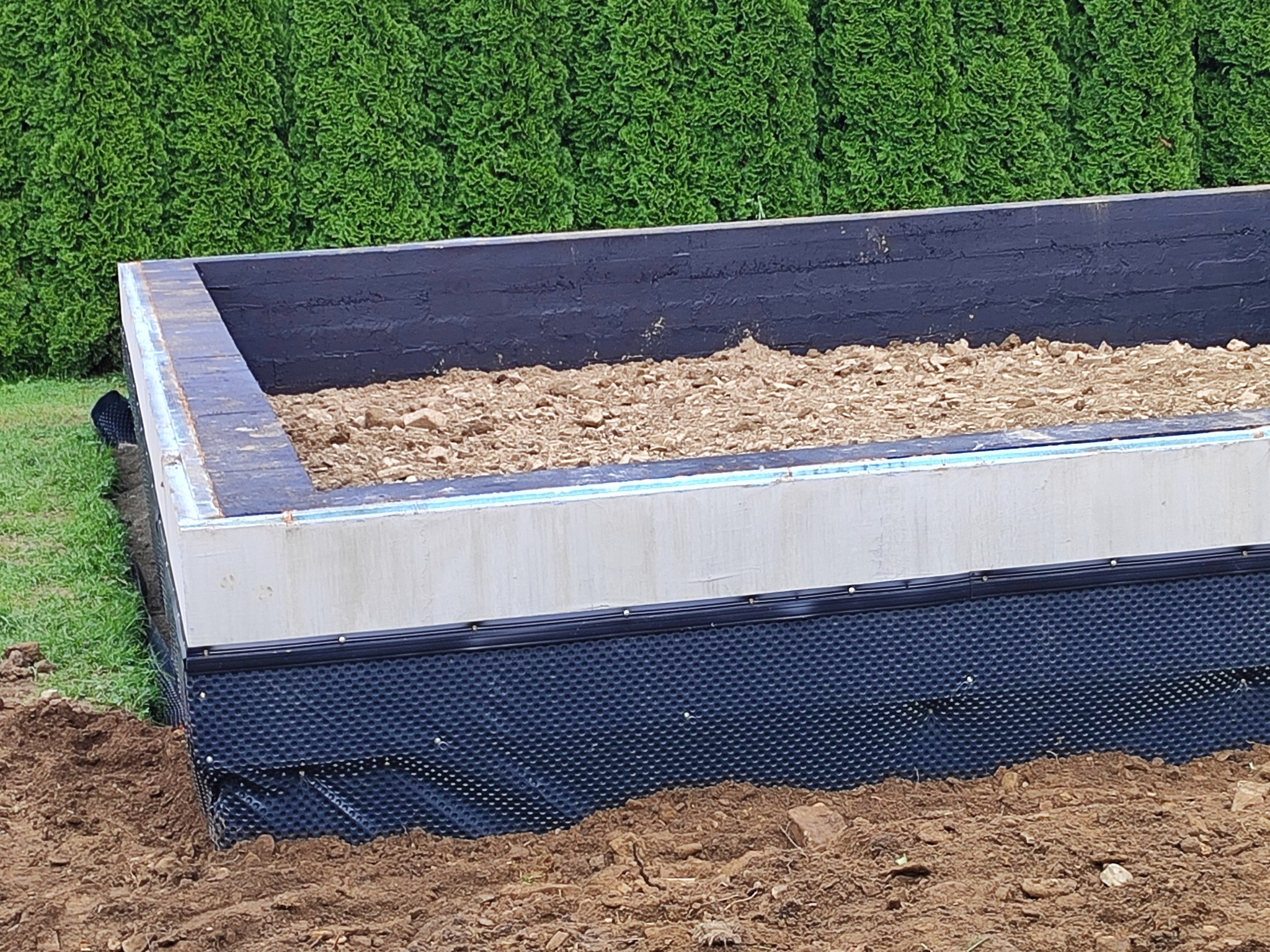 Fundament podwyższonej rabaty ogrodowej z izolacją pionową z folii kubełkowej, wypełniony ziemią i korą, na tle zielonych krzewów.