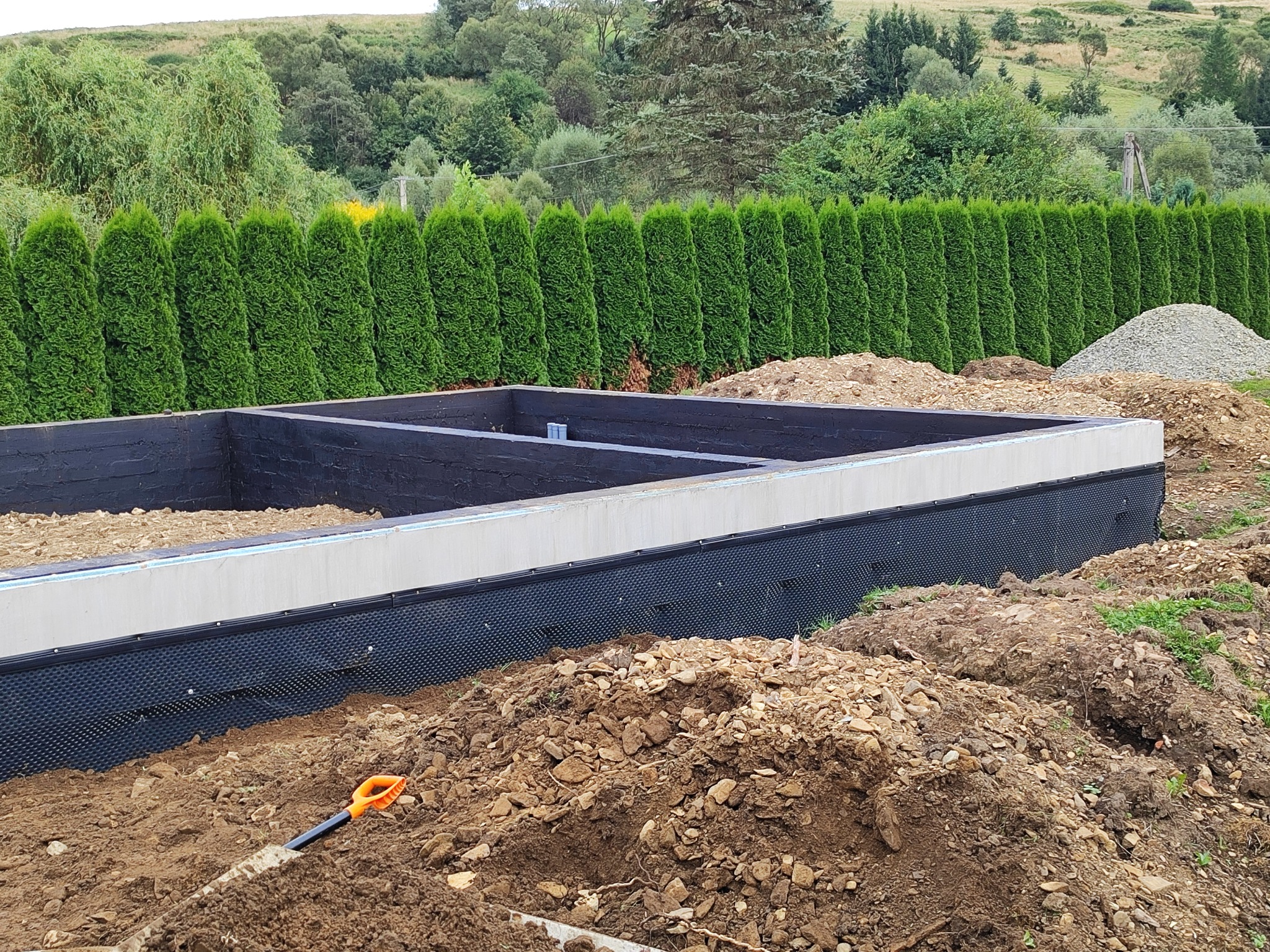 Wykop pod fundamenty budynku z izolacją przeciwwilgociową, widoczna łopata w ziemi i rząd tuj w tle.