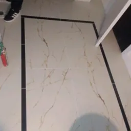 Ułożone płytki podłogowe w łazience, kremowe z brązowym marmurkowym wzorem, obramowane czarnymi paskami, widoczne fragmenty butów i puszka z pianką montażową.